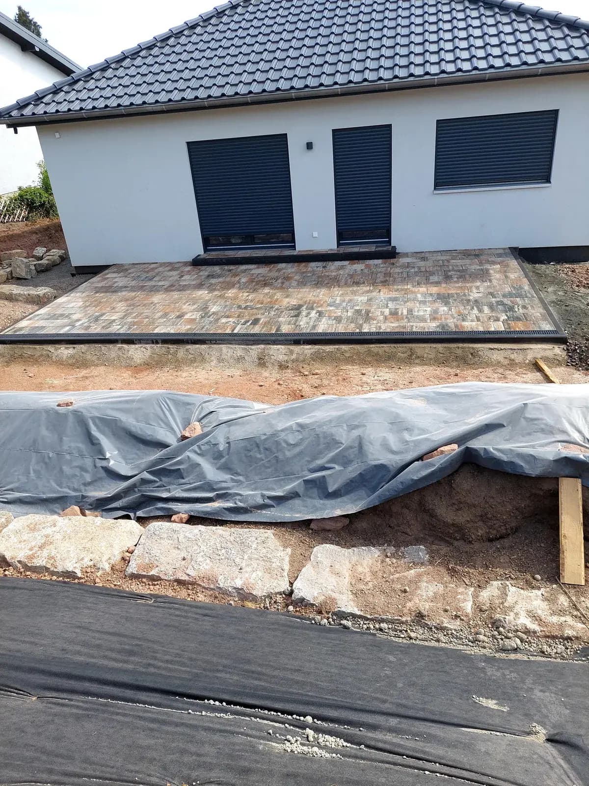 Nachher: fertig gepflasterte Terrassenfläche mit Natursteinmauer und modellierter Gartenfläche am Neubau
